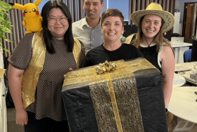 four people smiling holding large gold present box