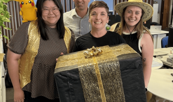 four people smiling holding large gold present box