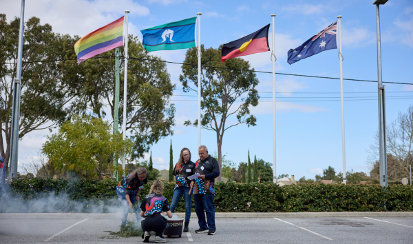 Four people around a fire with the rainbow Torres Strait Islander, Aboriginal and Australian flags behind them